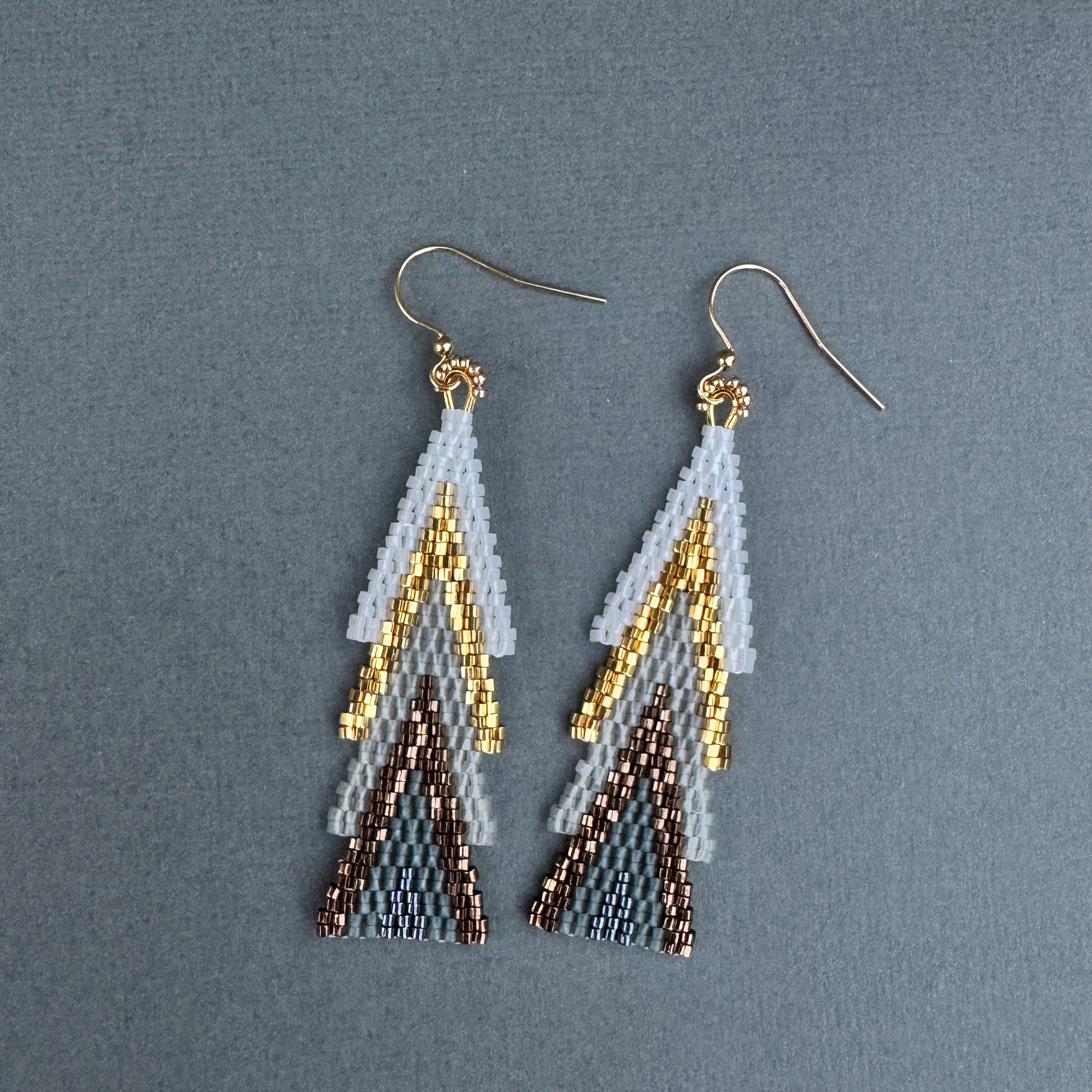 Beaded holiday earrings arranged flat, showing pine-inspired layers in white, gold, and bronze.”